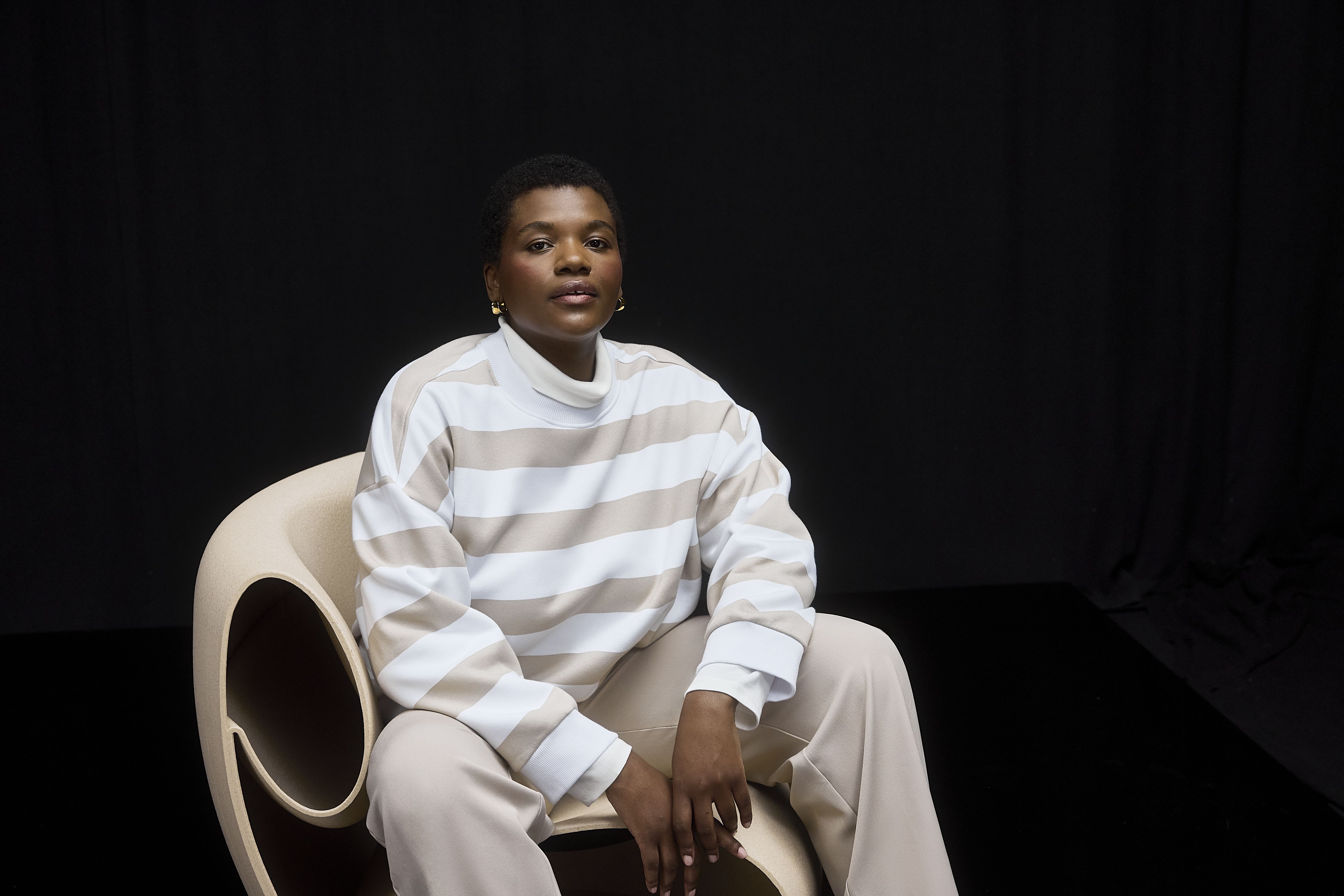 Mulher sentada, a usar uma camisola às riscas bege e branca, com calças bege e sapatilhas brancas