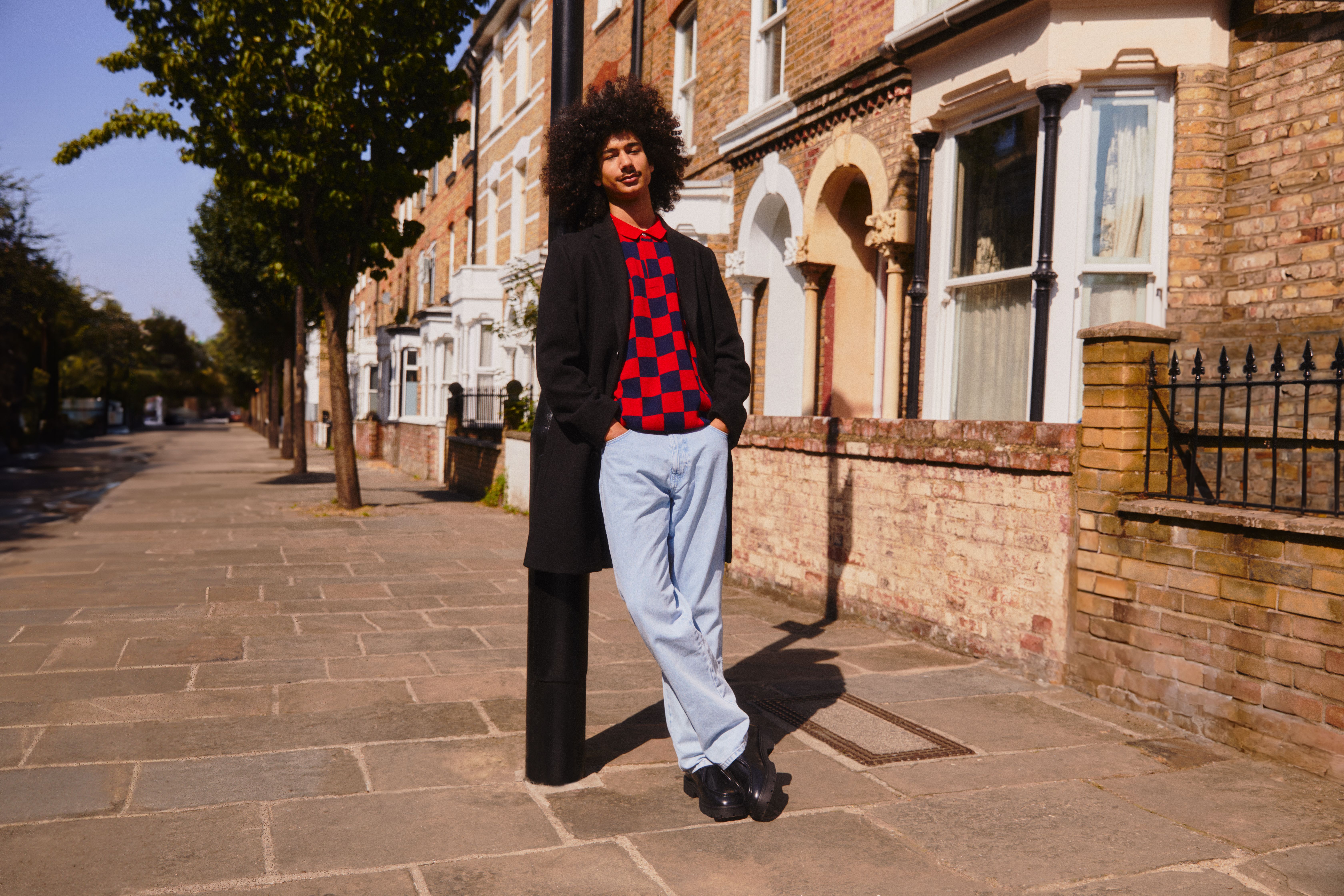 Homem encostado a um candeeiro de rua, a usar uma camisola vibrante e calças de ganga claras que se destacam no cenário